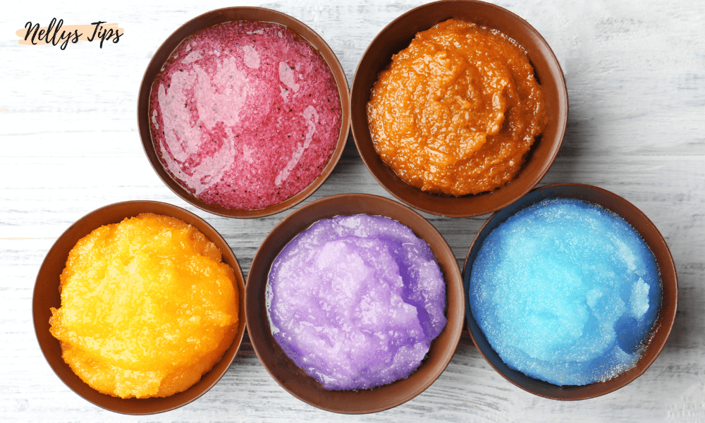 Fem skålar med olika färgade body scrubs: rosa, orange, gul, lila och blå, arrangerade på ett ljust träbord.