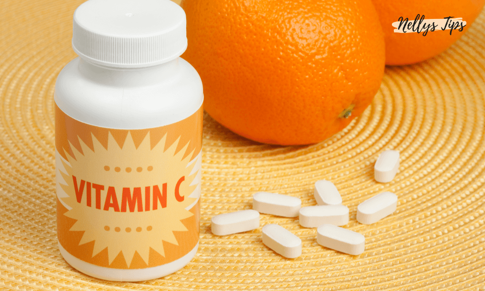 En flaska Vitamin C-tabletter bredvid några vita tabletter och två apelsiner på en guldfärgad yta. Flaskan har en gul etikett med röd text som säger 'VITAMIN C'.