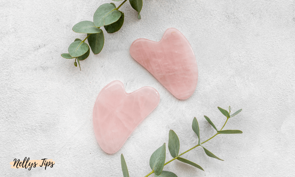 Två gua sha-stenar i rosékvarts placerade dekorativt med gröna blad på ljust underlag.