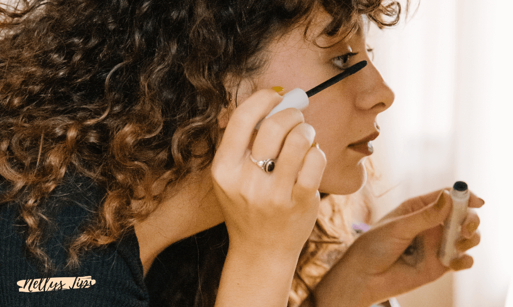 Kvinna med lockigt hår applicerar mascara framför spegel med vit borste