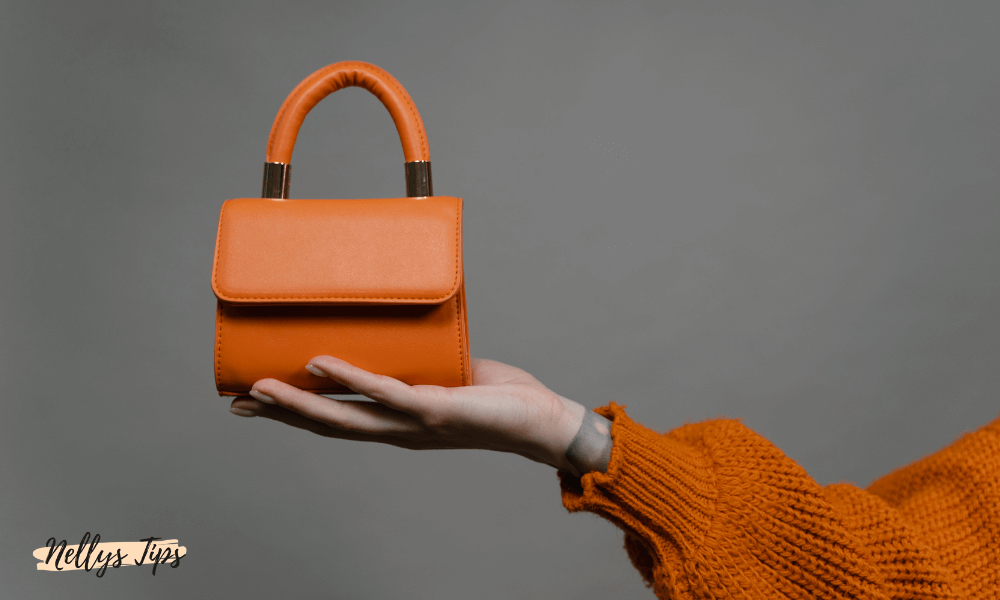 Kompakt orange handväska med rundat handtag, hållen i en hand med orange stickad ärm.