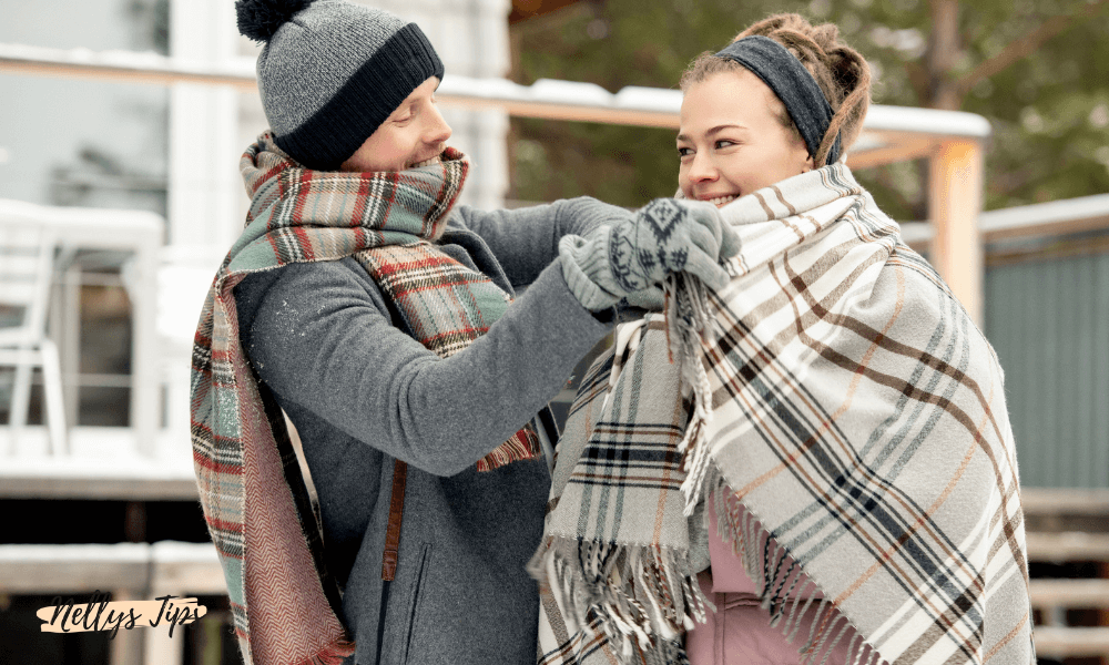 Man sveper en rutig halsduk runt en leende kvinna med filt, båda klädda för vinter.