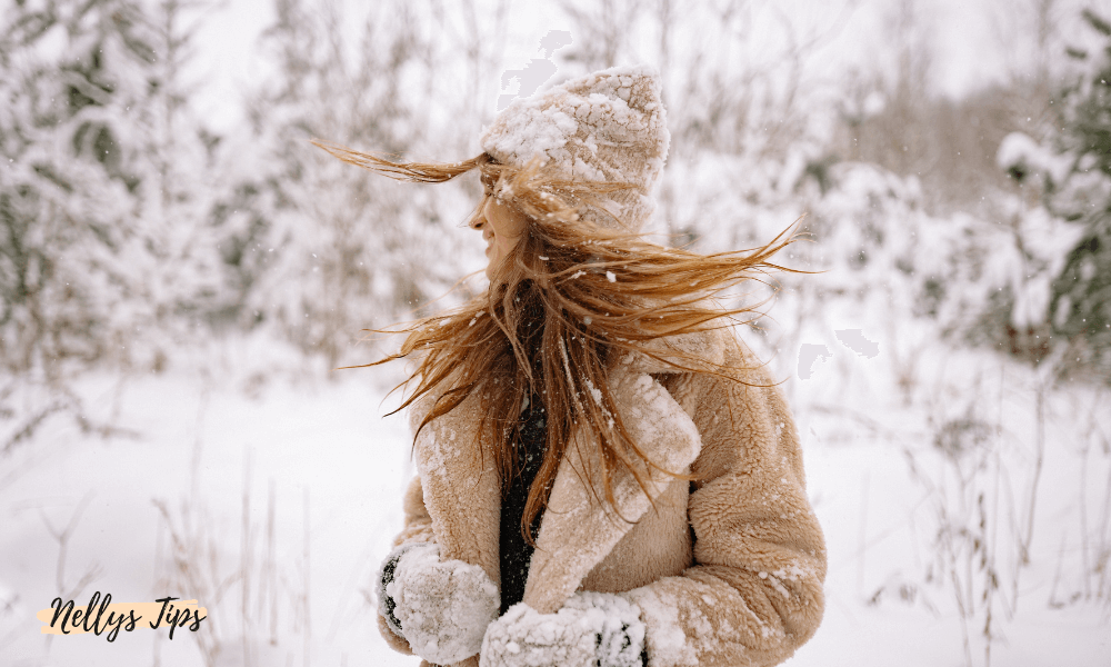 Kvinna i beige vinterjacka och stickad mössa i snöigt landskap, hår som blåser i vinden.