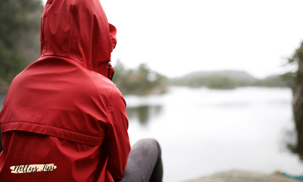 Kvinna i röd regnjacka med huva sitter vänd mot vattnet under en mulen dag.